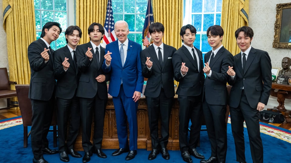 Sieben koreanische junge Männer in schwarzen Anzügen, links drei, rechts vier, in der Mitte alter Mann in blauem Anzug.