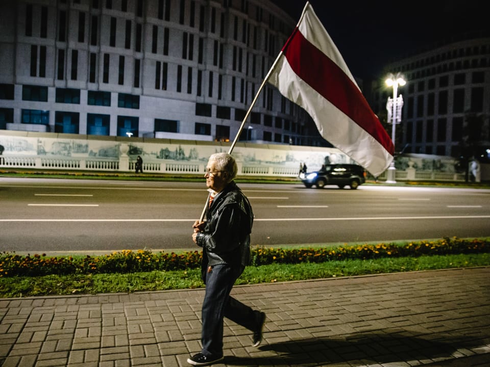 Ein älterer Mann läuft mit einer Flagge alleine die Strasse entlang.