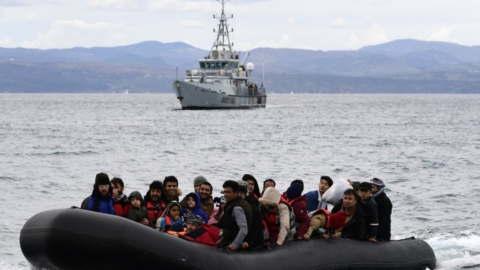 Boot mit Geflüchteten mit einem Schiff der Grenzschutzagentur Frontex im Hintergrund.
