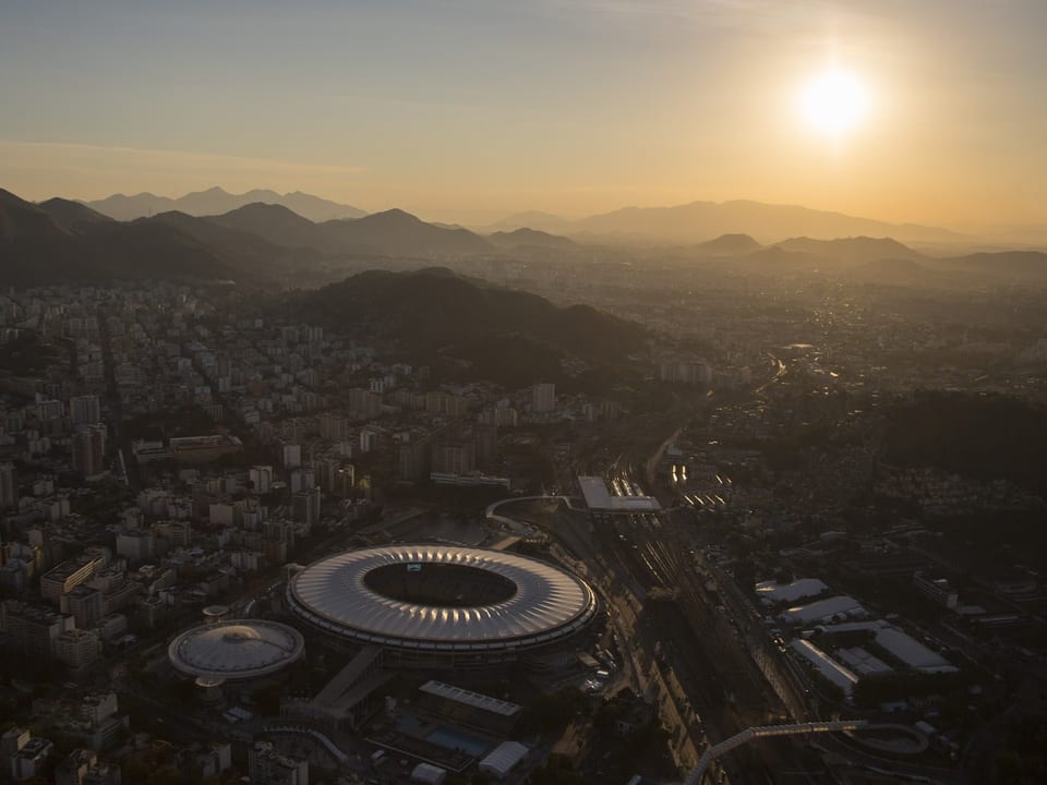 Luftaufnahme eines Stadions bei Sonnenuntergang in einer Stadt mit Hügeln im Hintergrund.