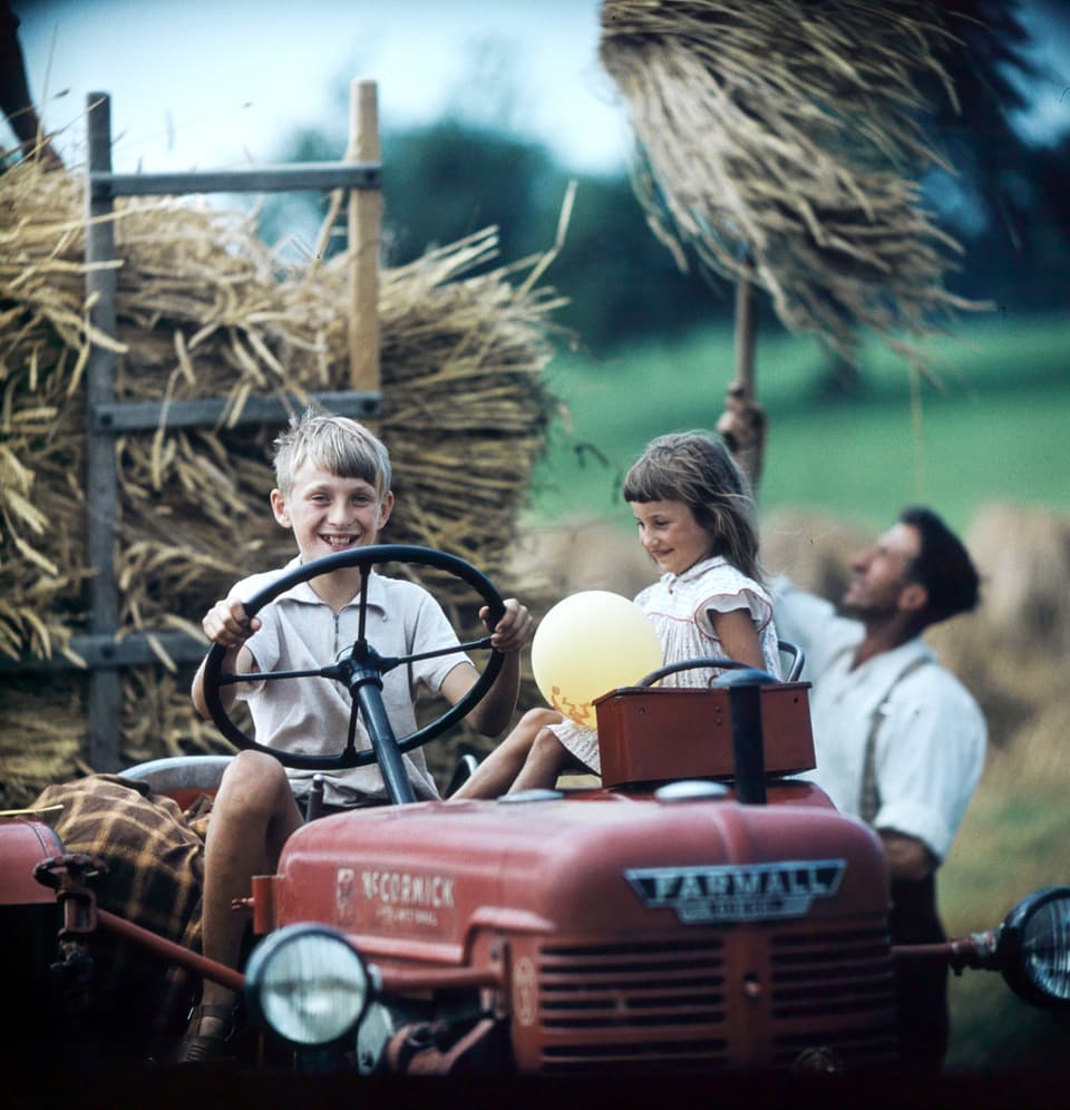 Ein Junge sitzt fährt lachend einen Traktor, neben ihm sitzend ein lachendes Mädchen, dahinter ein Mann, der Getreide mit einer Heugabel auf den Traktor schichtet.