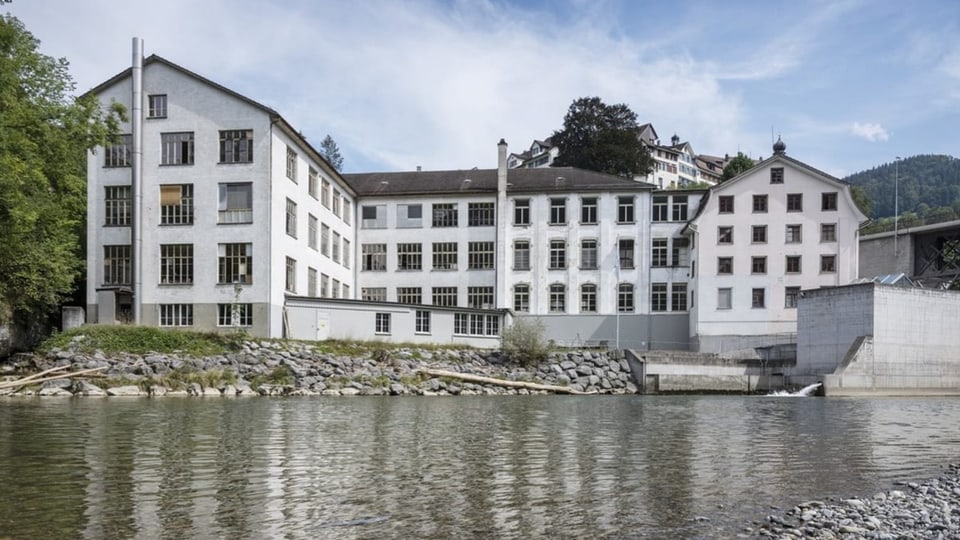 Ein umgenutztes viergeschossiges, weisses Fabrikgebäude mit grossen Fenstern am Flussufer.