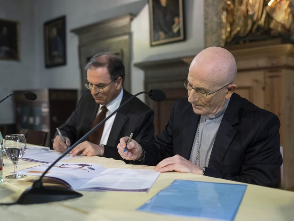 Ein Mann im Anzug und ein Mann im Bischofsgewand unterschreiben im Sitzen ein Dokument