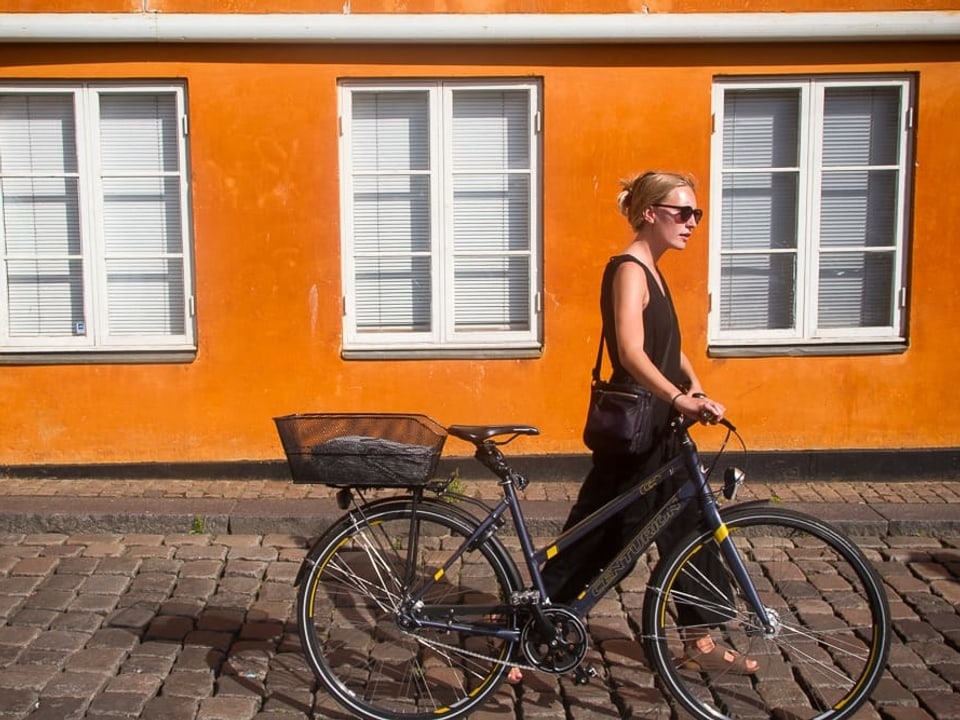 Eine junge Frau schiebt ihr Fahrrad an einer orangen Häuserfassade entlang.