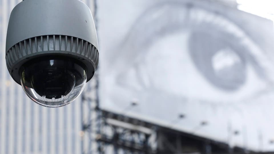 Eine Überwachungskamera, dahinter ein Plakat mit der Grossaufnahme eines Auges.