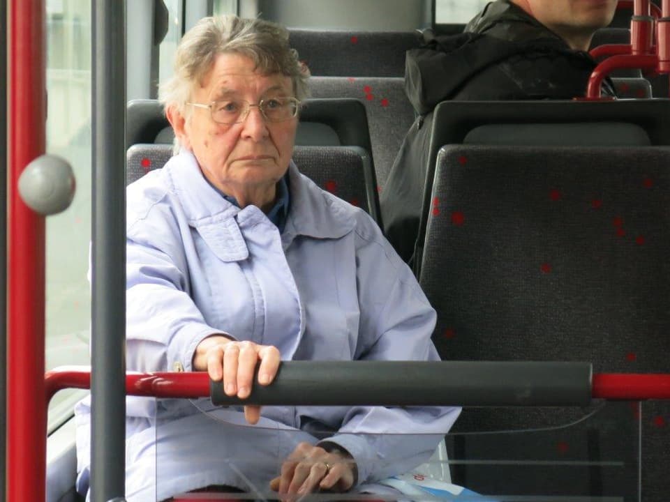 Eine ältere Frau sitz im Bus und hält sich an einer Stange fest.