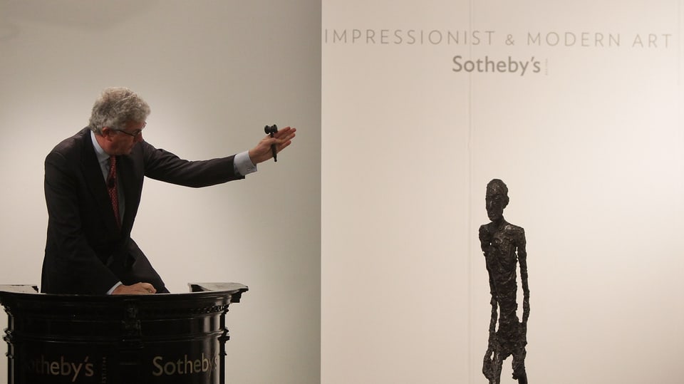 Ein Mann in Anzug und einem Hämmerchen in der Hand richtet seinen Arm auf eine Metallskulptur zu seiner Linken.