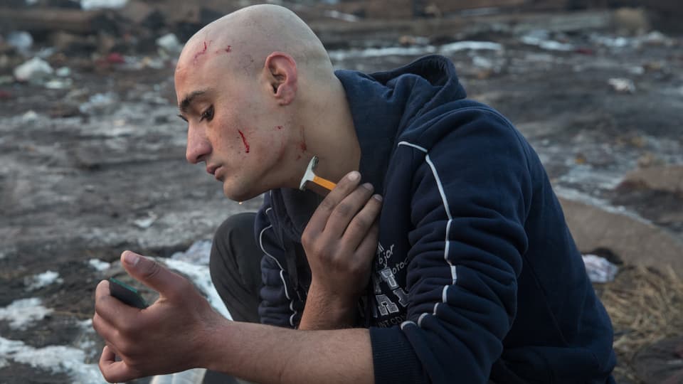 Ein Flüchtling mit kahlrasiertem Kopf rasiert sich draussen, mit einer Glasscherbe als Spiegel. Er hat sich an mehreren Stellen geschnitten und blutet.