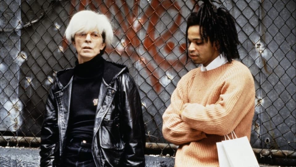 Andy Warhol und Basquiat stehen vor einem Maschendrahtzaun.