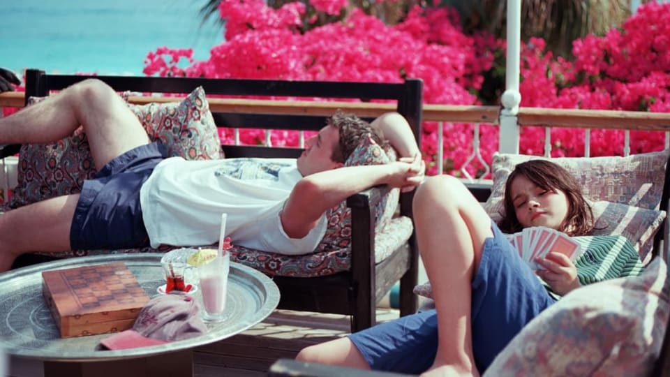 Mann und Mädchen liegen in Shorts auf Liegen auf einer Terrasse mit Meerblick.