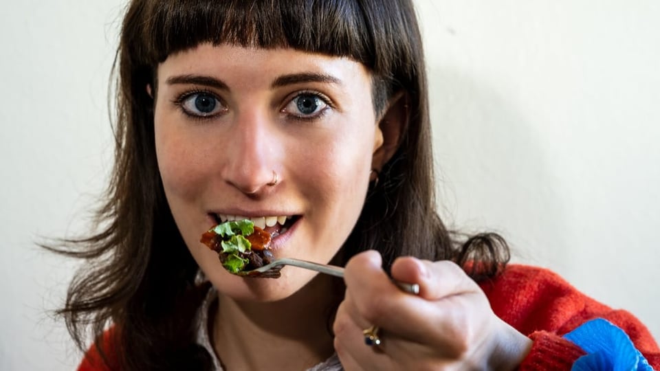 Junge Frau mit dunklen Haaren schaut in die Kamera, hält Gabel mit Salat vor dem Mund.
