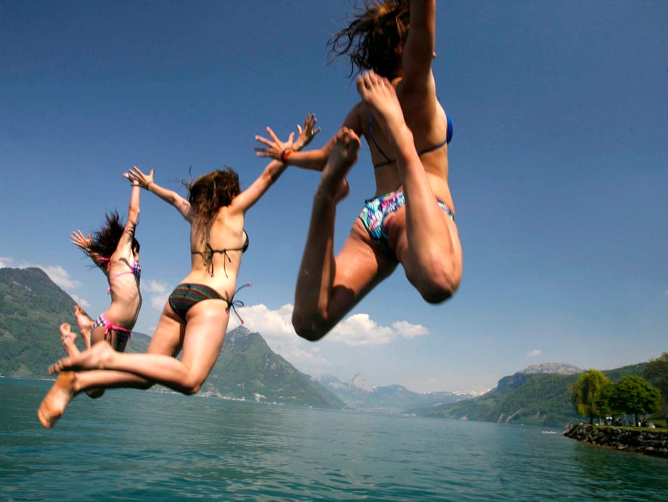 See bei sonnigem Wetter, im Hintergrund Ufer mit Bergen. Im Vordergrund drei Mädchen im Bikini, die ins Wasser springen. 