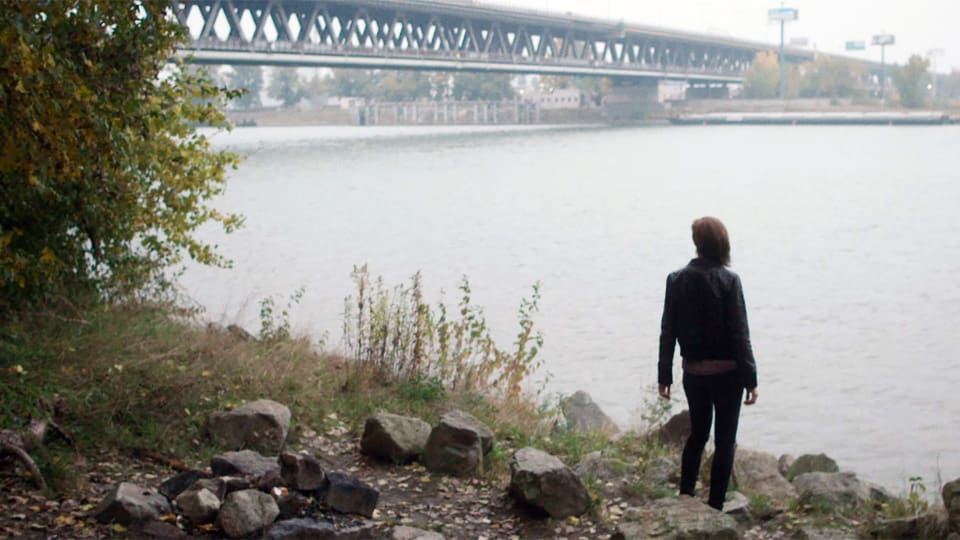 Eine Frau steht neben einer Brücke am Ufer eines breiten Flusses.