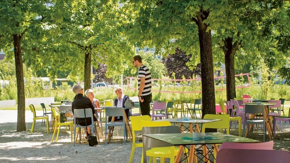 Menschen sitzen in einem grünen Park mit Tischen und Bäumen in Sitten.