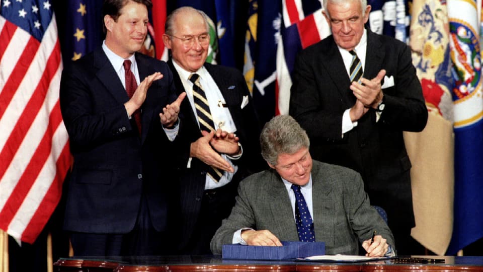 Bill Clinton unterzeichnet ein Abkommen, hinter ihm Vize-Präsident Al Gore und zwei andere Männer.