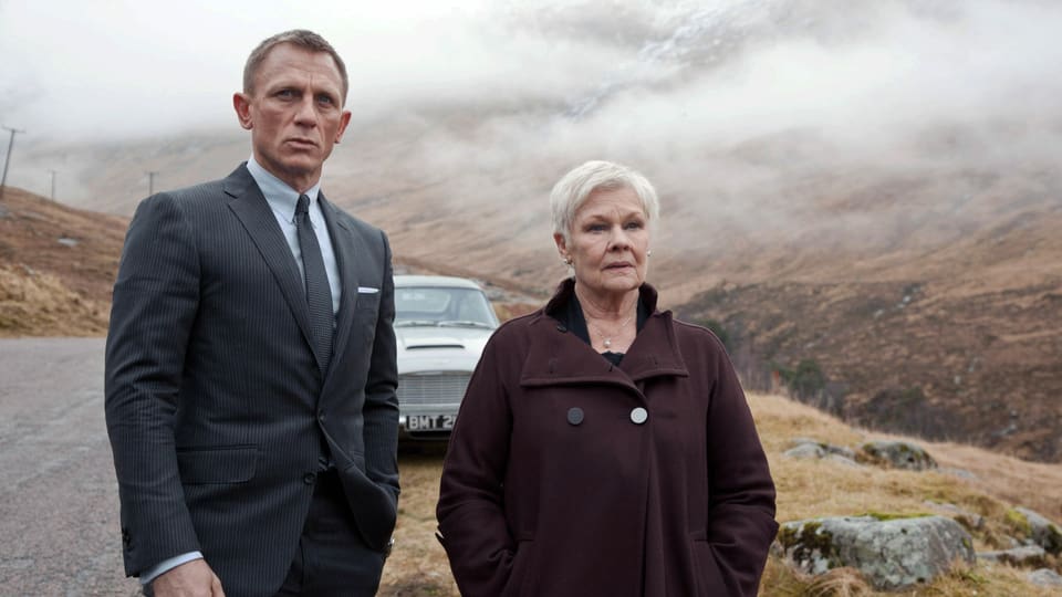 Ein Mann im Anzug und eine ältere Frau im Mantel stehen vor einem Auto in einer nebligen Landschaft.