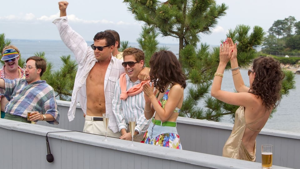 DiCaprio mit offenem Hemd, geballter Siegerfaust umgeben von Sekt, Bikini-Damen und Buddies.