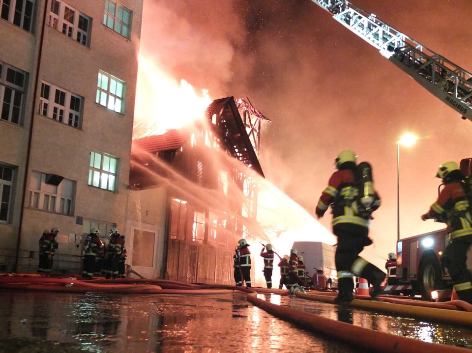Feuerwehrleute rennen umher, im Hintergrund ein grosses Gebäude in Vollbrand