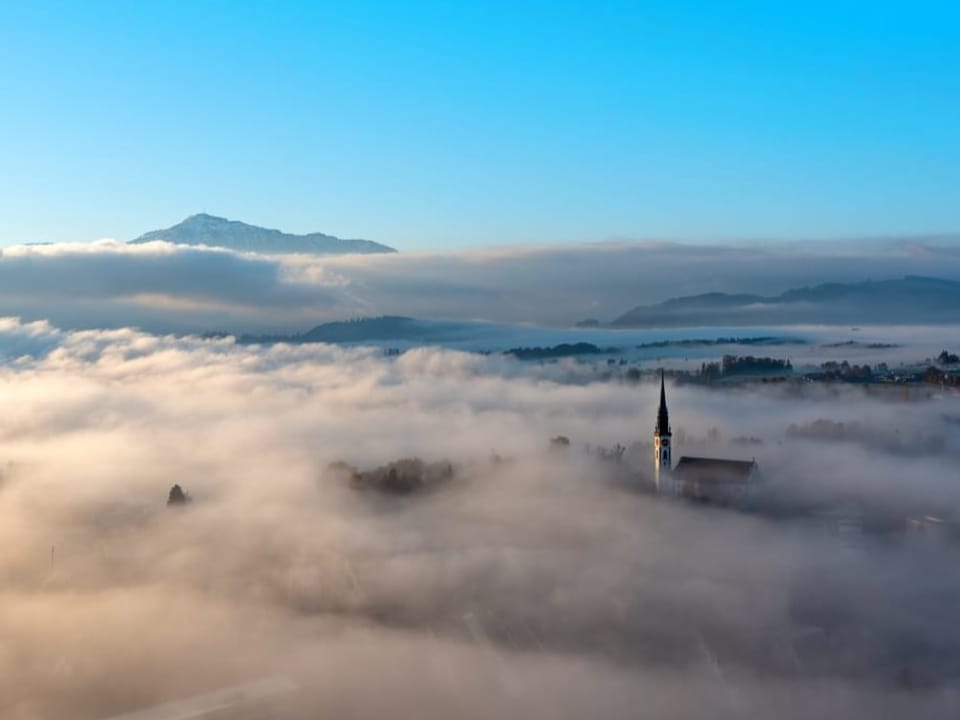 Luftbild auf Landschaft mit viel Bodennebel. Der Kirchturm eines Dorfs ragt aus dem Nebel. 