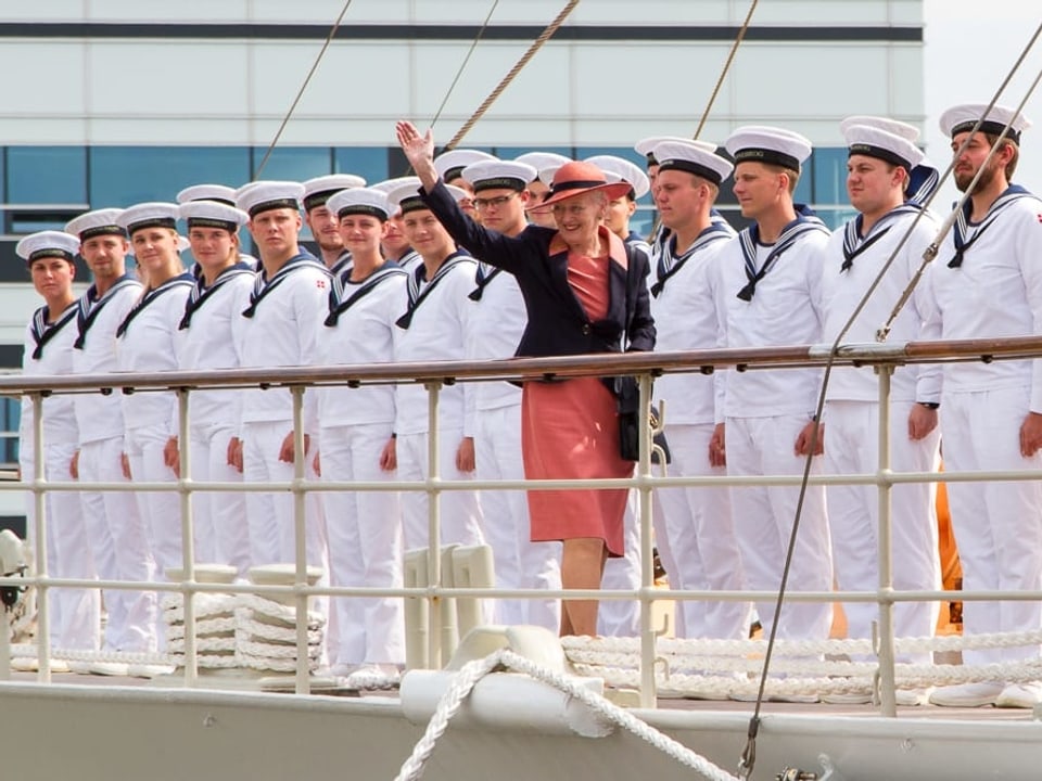 Königin Margrethe winkt von einem Schiff runter. Hinter ihr stehen Matrosen stramm.