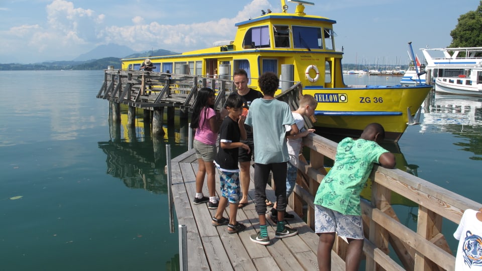 Ein schwimmendes Lagerhaus: Die MS Yellow auf dem Zugersee Eine Gruppe von Kindern steht auf einem Schiffssteg vor einem gelben Schiff.