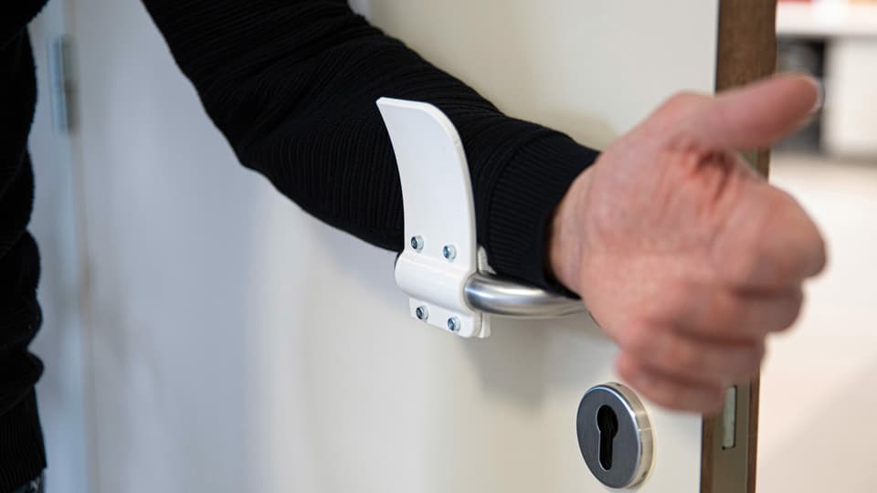 An einer Türfalle ist ein Plastikverlängerung angebracht, an der jemand mit dem Arm zieht.