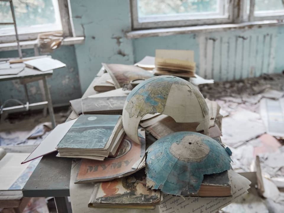 Ein kaputter Globus und alte Schulbücher liegen auf einem Tisch in einem verlassenen Schulzimmer.