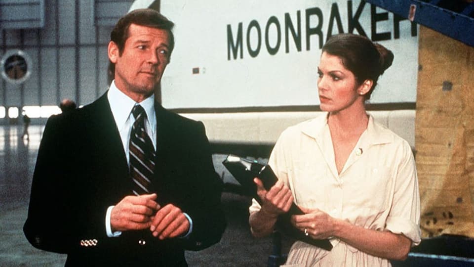 Ein Mann im Anzug steht neben einer Frau, dien Clipboard in der Hand hält – dahinter der Schriftzug «Moonraker».