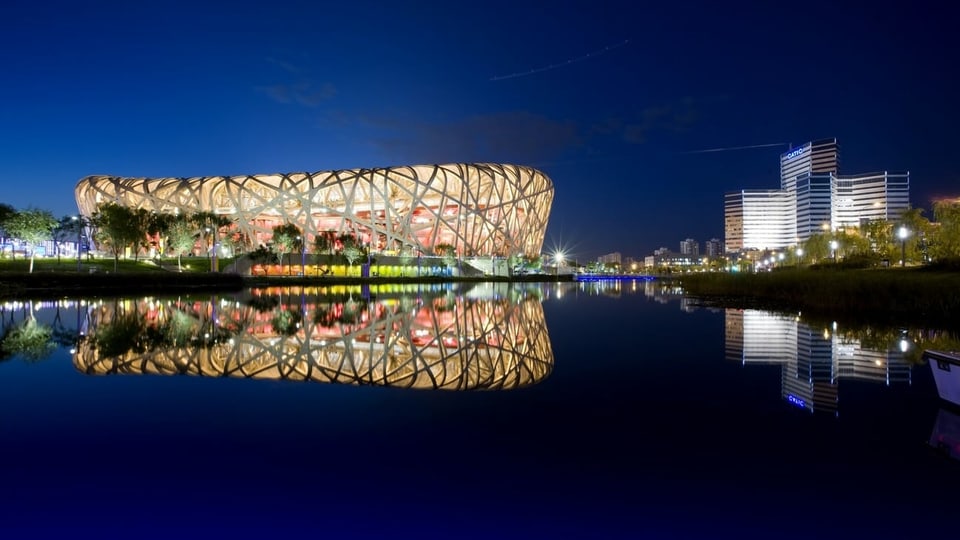 Sportstadion, das an ein Vogelnest erinnert, beleuchtet, in der Nacht, gespiegelt in einem Gewässer