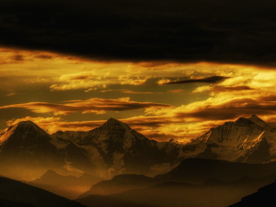 Gelb beleuchtete Alpen, darüber eine bedrohlöich schwarze Wolke.