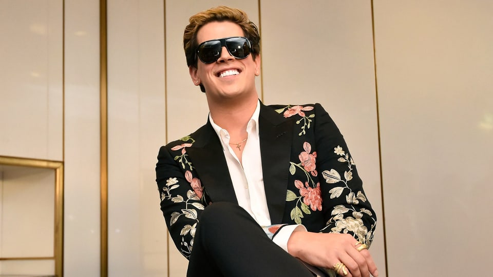 Milo sitzt mit einem goldbesticktem Sakko und vielen goldenen Ringen an den Fingern auf einem Stuhl.