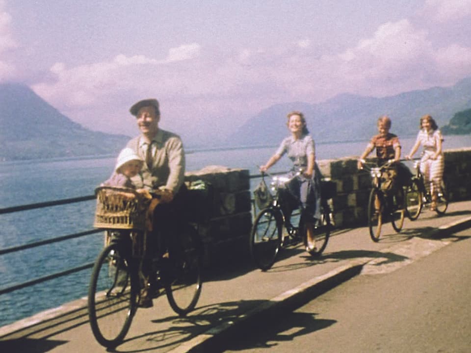 Filmsezene: Vier Menschen auf Fahrrädern fahren hintereinander einem Seeufer entlang.