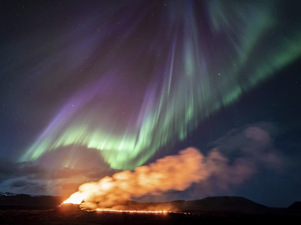 Nordlichter über einem Vulkan mit ausbrechender Lava und Rauch bei Nacht.