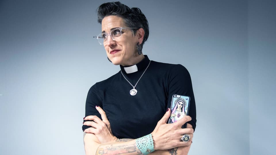 Eine Frau mit Kurzhaarschnitt und Tattoos hält ein Smartphone in der Hand. Sie ist Pfarrerin und trägt ein schwarzes T-Shirt, am Hals ist eine weisse Pfarrersbinde und eine Goldkette.
