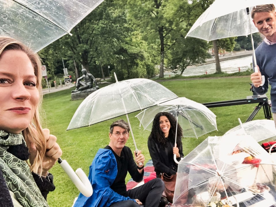 Franz Fischlin, Andrea Jansen, Peter Brönnimann und Mujinga Kambundji mit Regenschirmen sitzen auf einer Picknick-Decke in der Wiese.