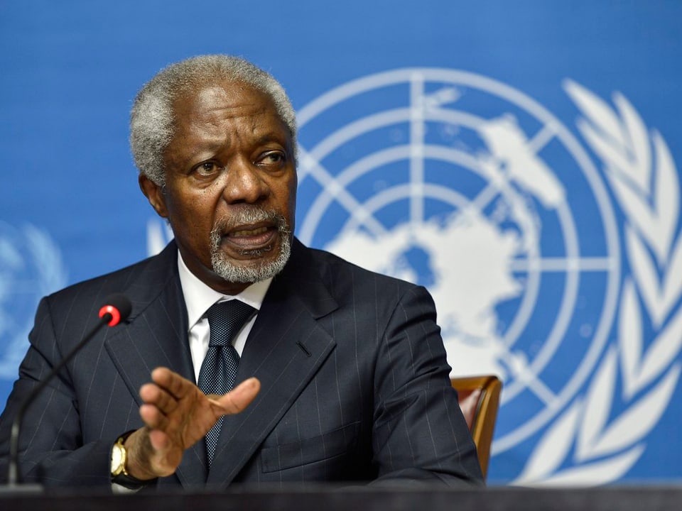 Kofi Annan sitzt vor einem Mikrofon. Im Hintergrund das Logo der UNO.