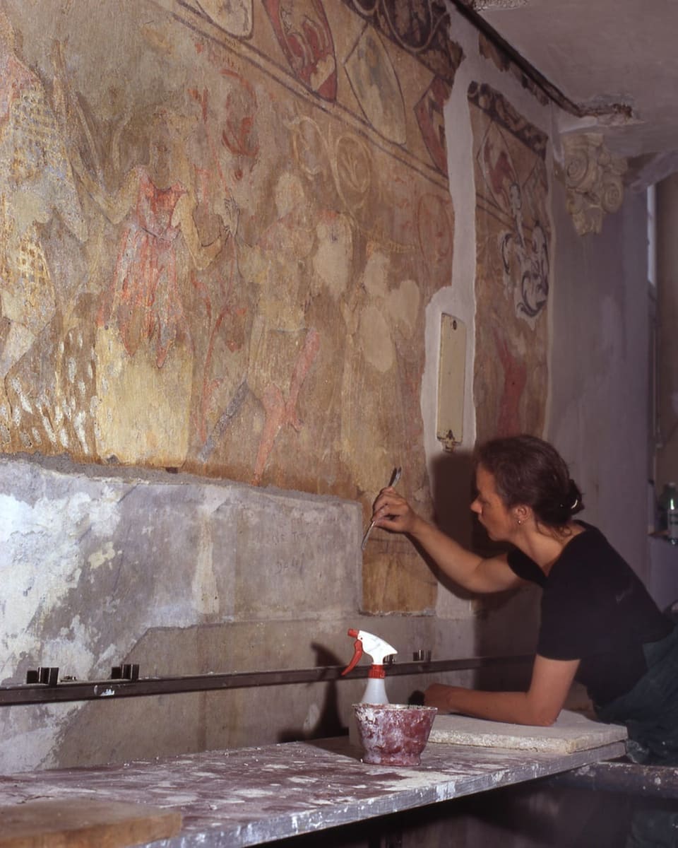 Foto, links leicht orange Wandmalerei, rechts eine Frau mit Werkzeug, auf Wand konzentriert.