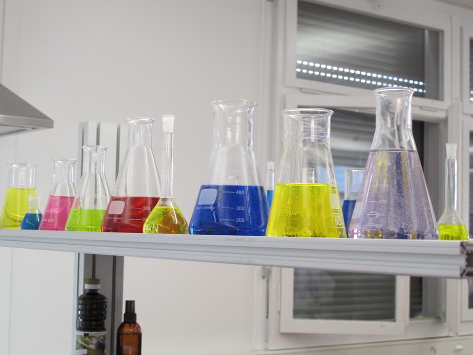 Auf einem Laborregal stehen Glasbehälter mit farbigen Flüssigkeiten.