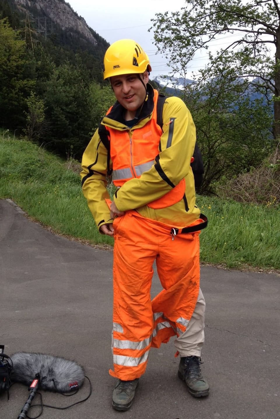 Mann mit gelbem Helm, gelber Jacke und Leuchtweste zieht sich leuchtend orange Hosen an.