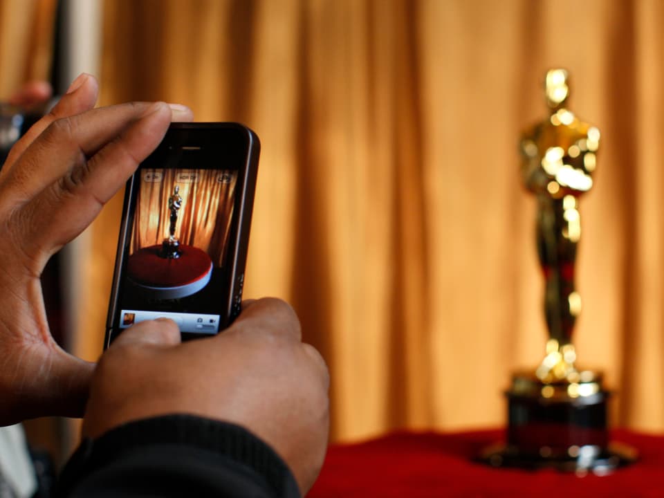 Zwei Hände halten ein Mobiltelefon, mit dem ein Foto einer goldenen Oscar-Statue geknippst wird.