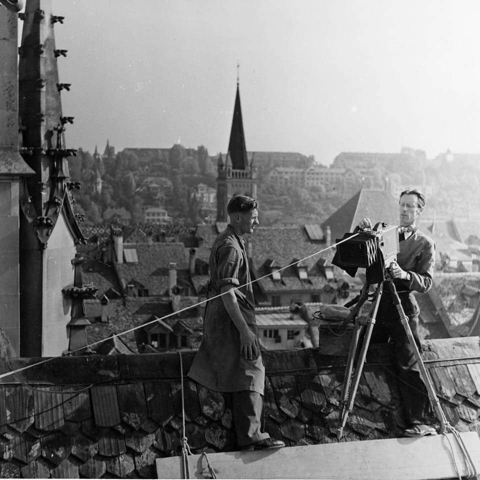 Der Kameramann und sein Assistent stehen mit der Kamera, welche mit Seilen befestigt ist, auf dem Dach der Berner Münsters.