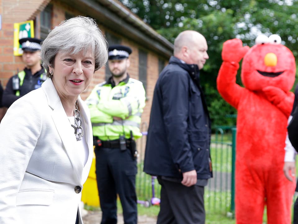 Theresa May im Vordergrund, im Hintergrund Polizisten und ein winkender Mann in rotem Elmo-Kostüm.