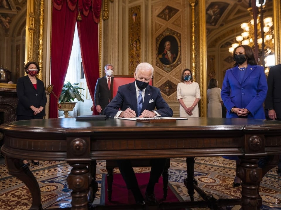 Joe Biden sitzt mit einer Maske an einem Tisch und unterzeichnet ein Dokument.