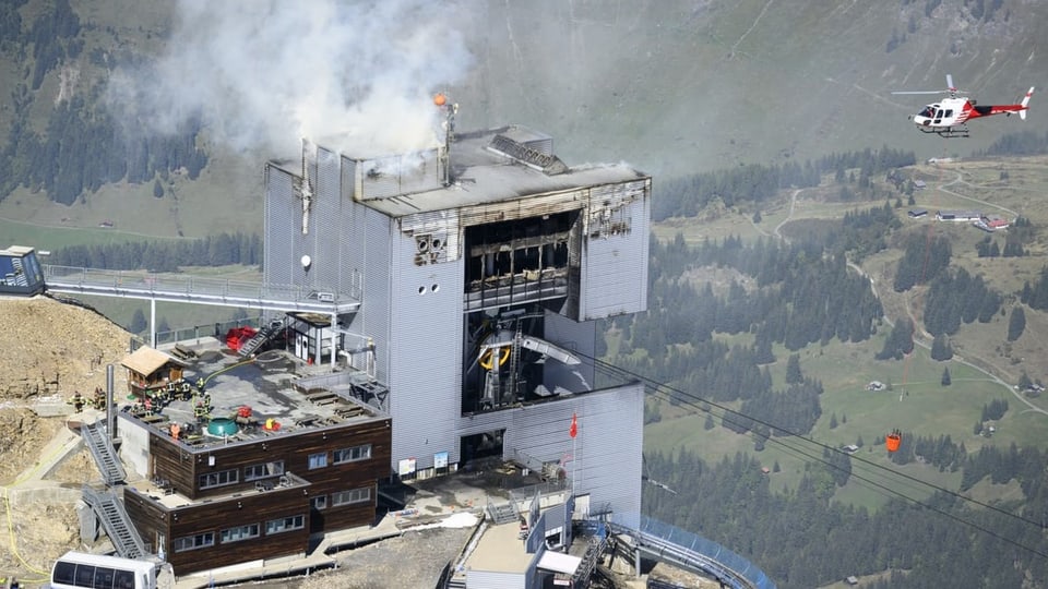 Ein Löschhelikopter fliegt Richtung Bergstation einer Drahtseilbahn, aus welcher Rauch aufsteigt.