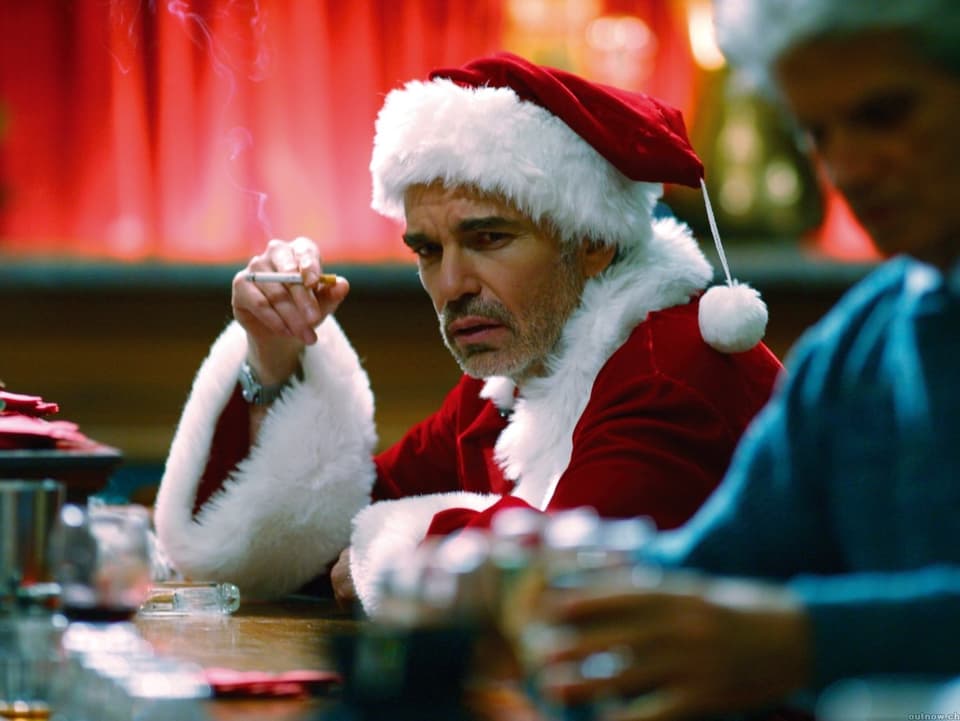 Billy Bob Thornton im Weihnachtsmann-Kostüm, er sitzt schlecht gelaunt und rauchend an einer Bar.