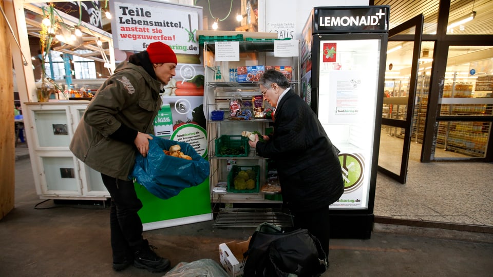 Ein junger Mann und eine ältere Frau stehen vor einem offenen Kühlschrank, der vor einem Supermarkt platziert ist.