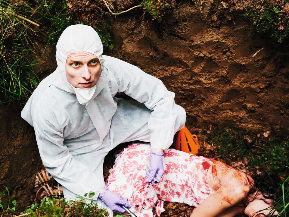 Ein Mann in weisser Schutzkleidung sitzt in einer Grube neben der Leiche einer Frau.