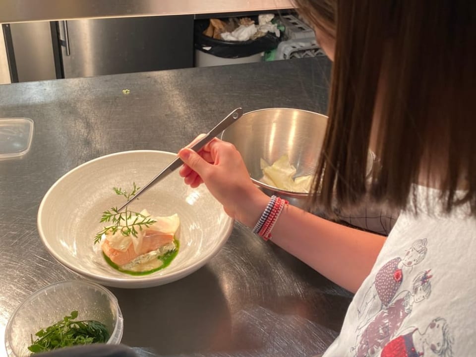 Auf einem Metalluntergrund steht ein Teller mit Fisch. Ein Mädchen hält eine Pinzette in der Hand.