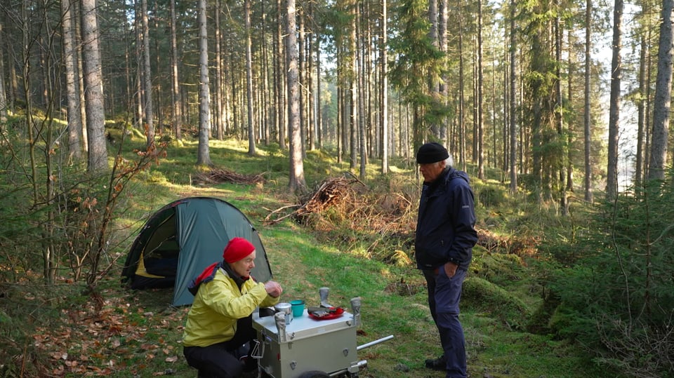 Zwei Männer essen im Wald, neben einem Zelt.