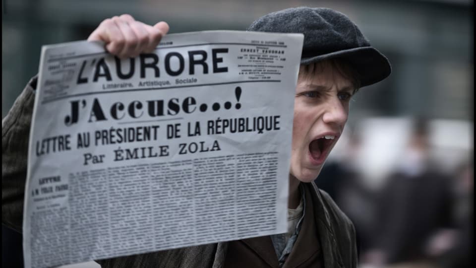 Zeitungsjunge hält schreiend eine Zeitunng in die Luft, auf der der Titel «J'accuse!» zu lesen ist.